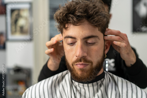 Making haircut look perfect. Young bearded man getting haircut by hairdresser while sitting in chair at barbershop - small business and people lifestyle concept