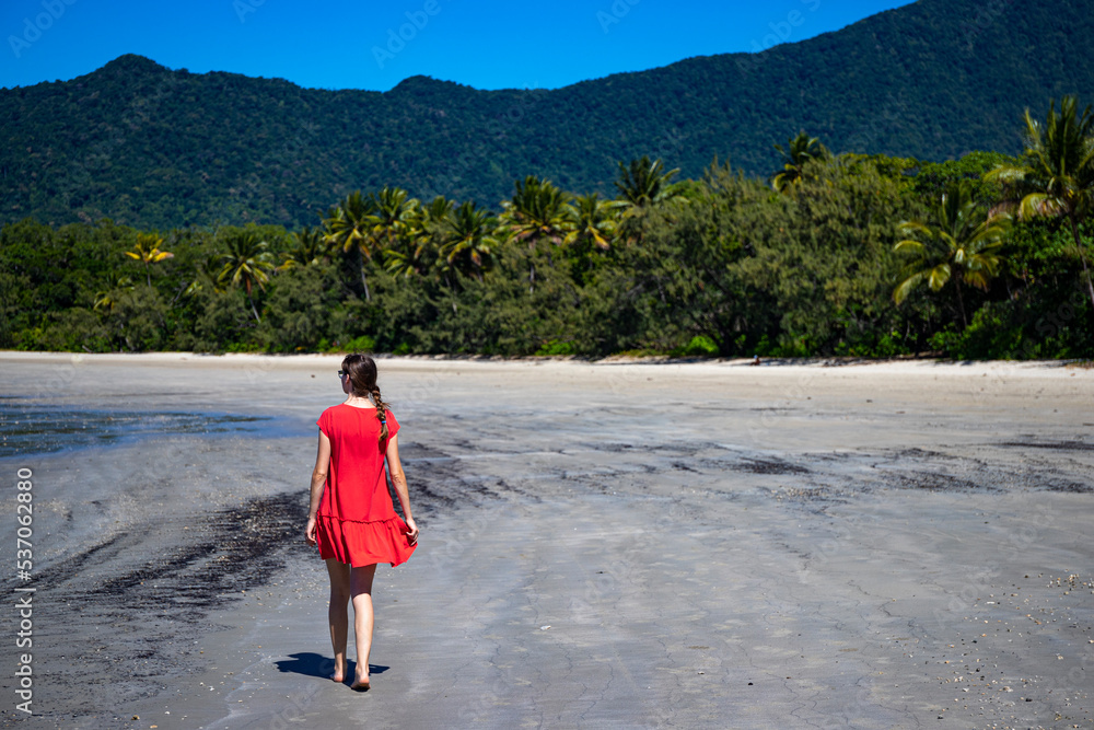 a beautiful girl in a red dress enjoys the sun on a tropical beach with palm trees in the background in daintree national park, vacation in queensland, australia; daintree rainforest
