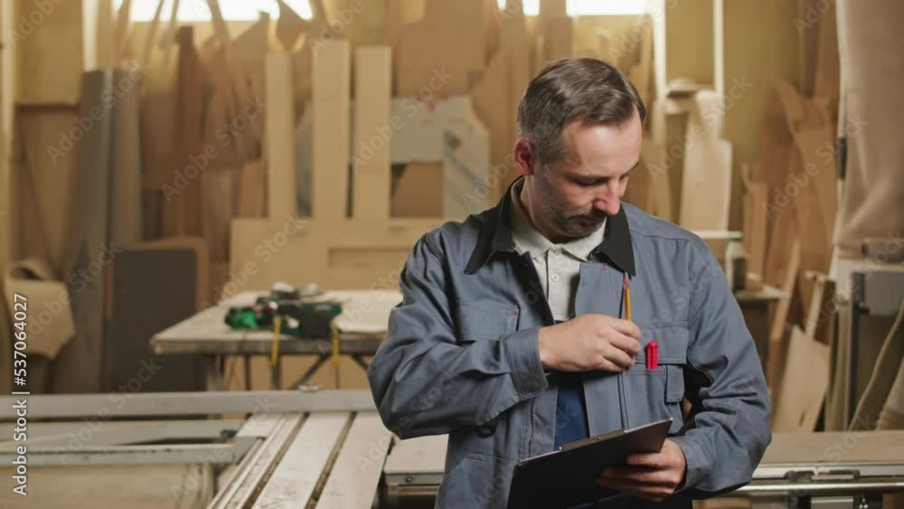 Medium slowmo portrait of middle aged Caucasian joiner taking notes on clipboard and smiling at camera in woodworking workshop Medium slowmo portrait of middle aged Caucasian joiner taking notes on clipboard and smiling at camera in woodworking workshop