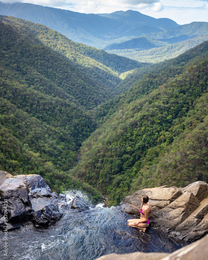beautiful girl in bikini sits over precipice at top of windin falls; scary waterfall in atherton tablelands, queensland, australia, pool at top of waterfall