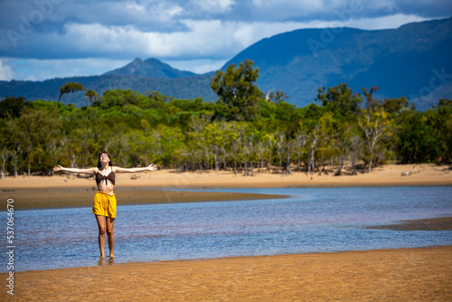 A beautiful girl in a bikini walks on a tropical Australian beach with palm trees and mighty mountains in the background; relaxing in northern queensland, a hot day at the beach