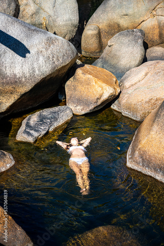 a beautiful girl in a white bikini lies on the water in a natural pool surrounded by massive rocks in jourama falls; relaxing in paluma range national park in queensland, australia; cascading waterfal photo