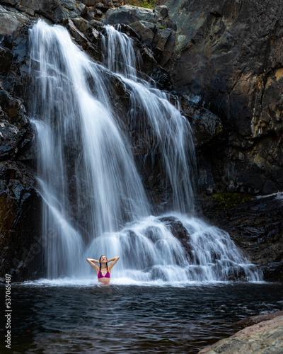 a beautiful girl in a pink bikini stands under a waterfall in a pool surrounded by massive rocks in jourama falls; vacation in paluma range national park in queensland, australia; cascading waterfalls