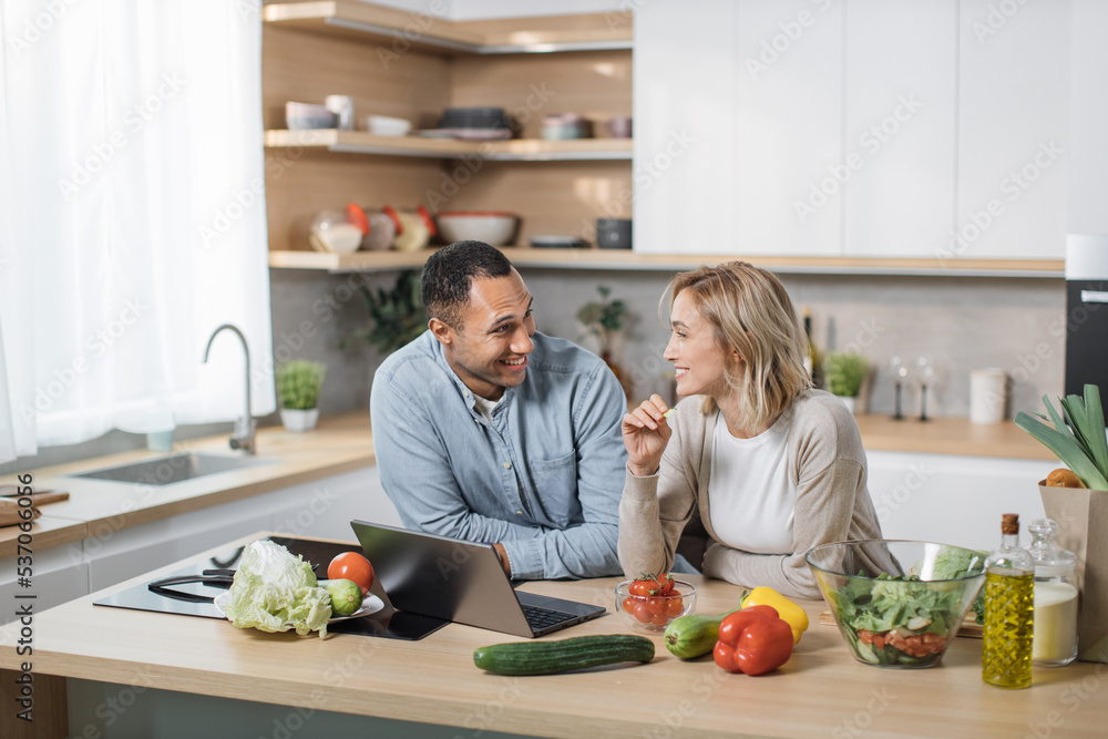 Happy young multiracial couple using laptop computer preparing healthy food diet vegetable salad at home together, searching recipes, ordering shopping online, watching cooking class in kitchen.