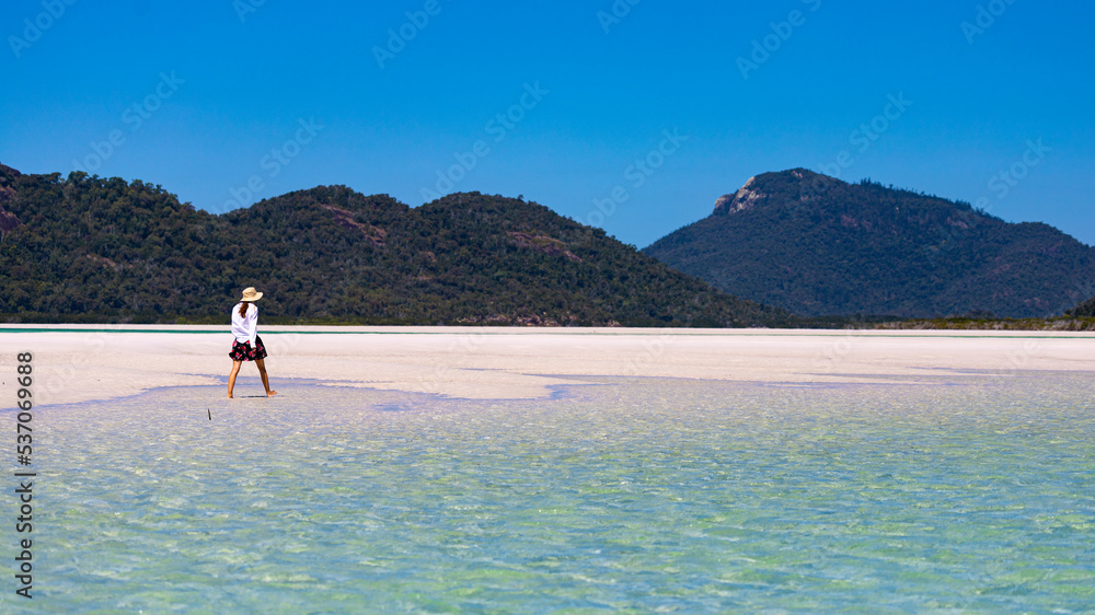 a beautiful girl in a dress, shirt and hat enjoys a walk on the white dunes at whitehaven beach; paradise beach with white sand and turquoise waters; whistundays island holiday, queensland, australia;