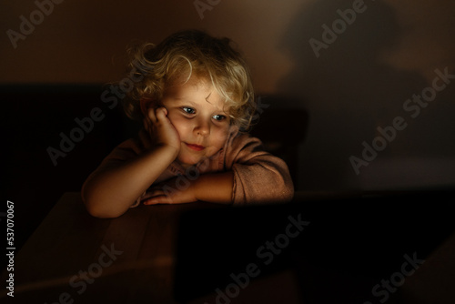 Portrait of a cute child. a girl watches a tablet with cartoons or a movie in the evening. Lamp light in the room.