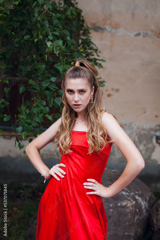 A cute young girl with blond flowing long hair and beautiful make-up, in a long red dress, stands on the street of the old city. There is a tree nearby. A cute young girl with blond flowing long hair and beautiful make-up, in a long red dress, stands on the street of the old city. There is a tree nearby.