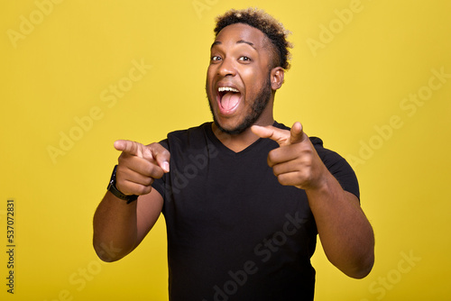 Dark-skinned smiling guy points forward in front of him with two hands into camera. Happy young man with a smartwatch points his index finger at the camera on a yellow background