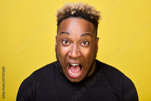An outraged African-American yells with all his might in outrage close-ups on camera on a yellow background. An enraged black guy screams loudly with his mouth wide open and bulging eyes.  © Aleksandr