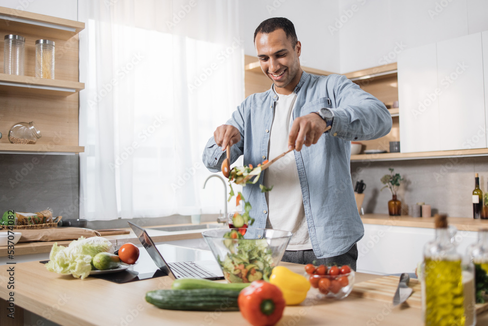 Obraz premium Focus on man's hands holding wooden spoons and mixing ingredients in a glass bowl, preparing delicious healthy breakfast or dinner in the modern kitchen island.