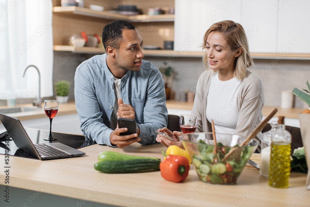 Happy multinational couple using cell phone while having breakfast at kitchen, positive man and woman eating healthy food, checking photos on social media and smiling, having video conference