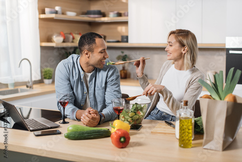 Cheerful married multinational couple using laptop while cooking healthy food in kitchen  blond young wife spouses having fun while feeding her husband wooden spoon with salad from fresh vegetables