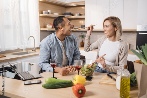 Cheerful married multinational couple using laptop while cooking healthy food in kitchen, blond young wife spouses having fun while feeding her husband wooden spoon with salad from fresh vegetables