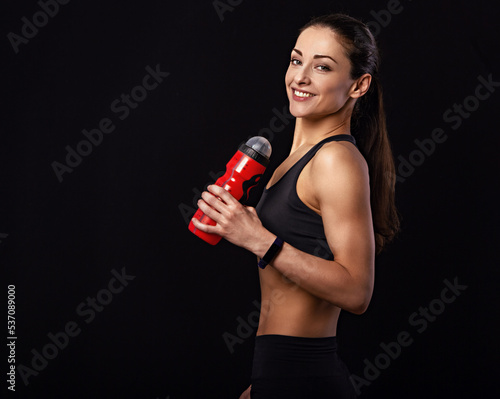 Happy pretty fit healthy fitness woman in  sporty top clothing holding sport flask and looking down on black background with empty copy space. Healthy