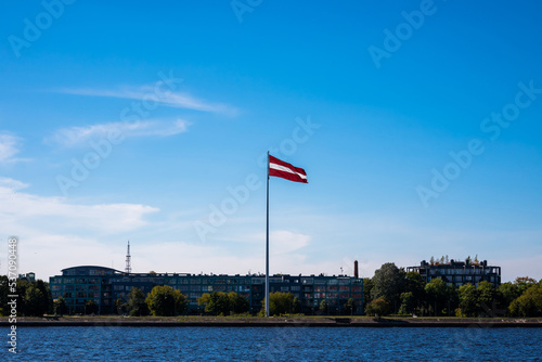 Latvian Flag Hang On Flagpole Against Blue Sky, On Banks Of Daugava River.