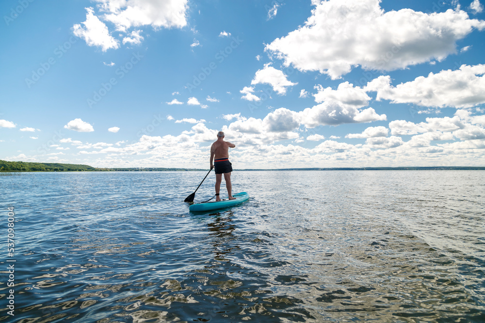 A man on a SUP board with a paddle in the lake on a sunny day against the backdrop of white clouds.