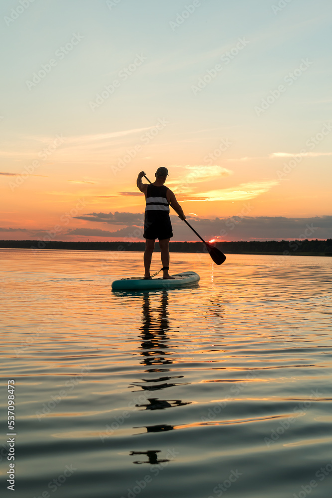 A man in shorts on a SUP board with an oar against the backdrop of a bright sunset sky swims in the lake in the evening.