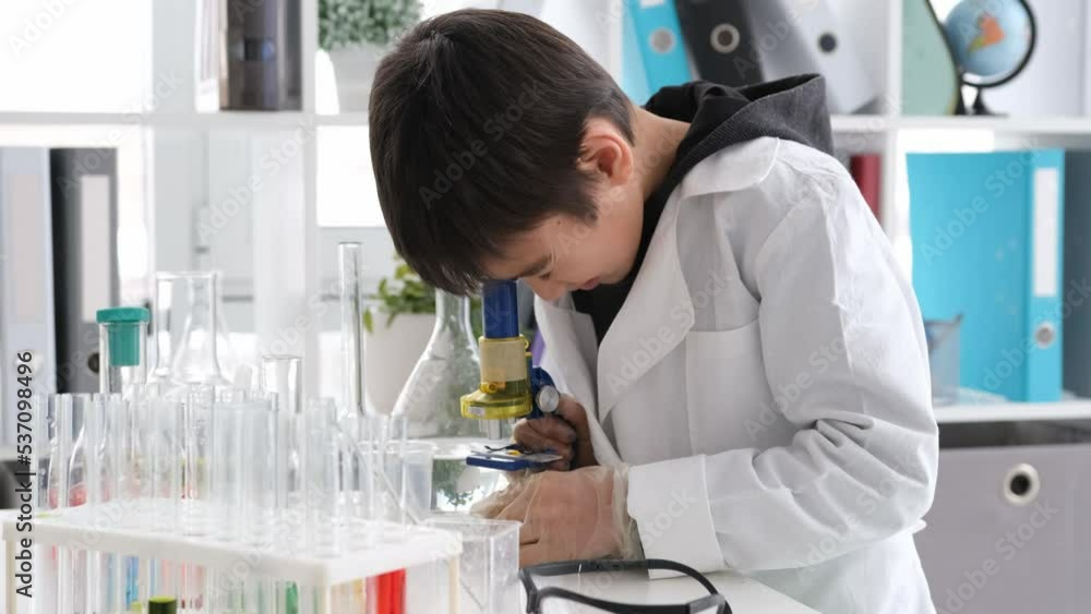 Schoolboy at chemistry lesson with microscope looking reaction at class with tubes and equipment for tests. Pupil boy in lab studying Schoolboy at chemistry lesson with microscope looking reaction at class with tubes and equipment for tests. Pupil boy in lab studying