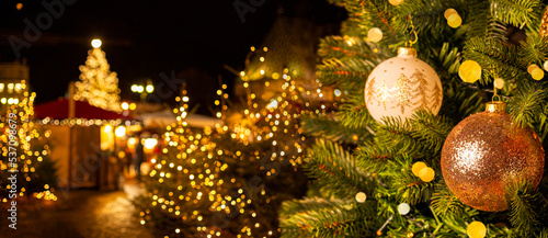 European Christmas city with festive fair or market in the evening. Christmas tree with beautiful balls on foreground. Holiday background.