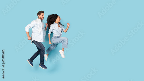 Happy millennial caucasian man and arab female fast run, jump in air to empty space, isolated on blue background