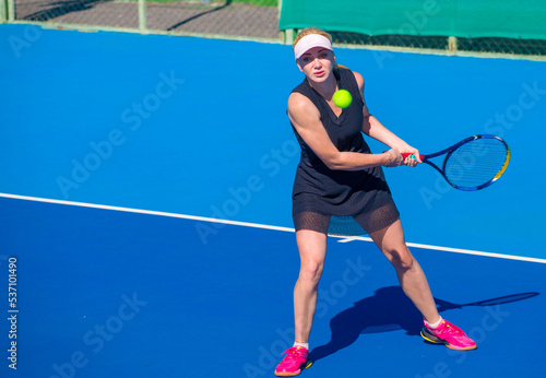 A girl plays tennis on a court with a hard blue surface on a summer sunny day