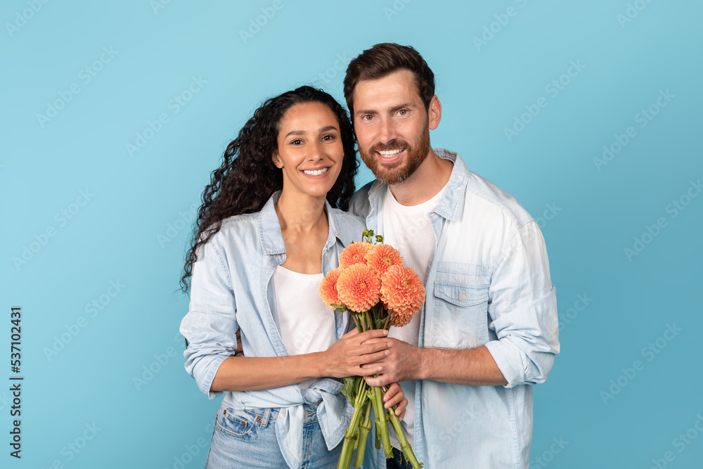 Glad millennial arab female and european man with beard looking at camera and holding flowers bouquet