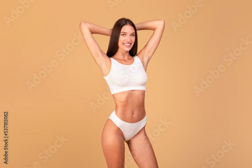 Attractive fit woman wearing white lingerie and posing with hands behind head isolated on beige studio background