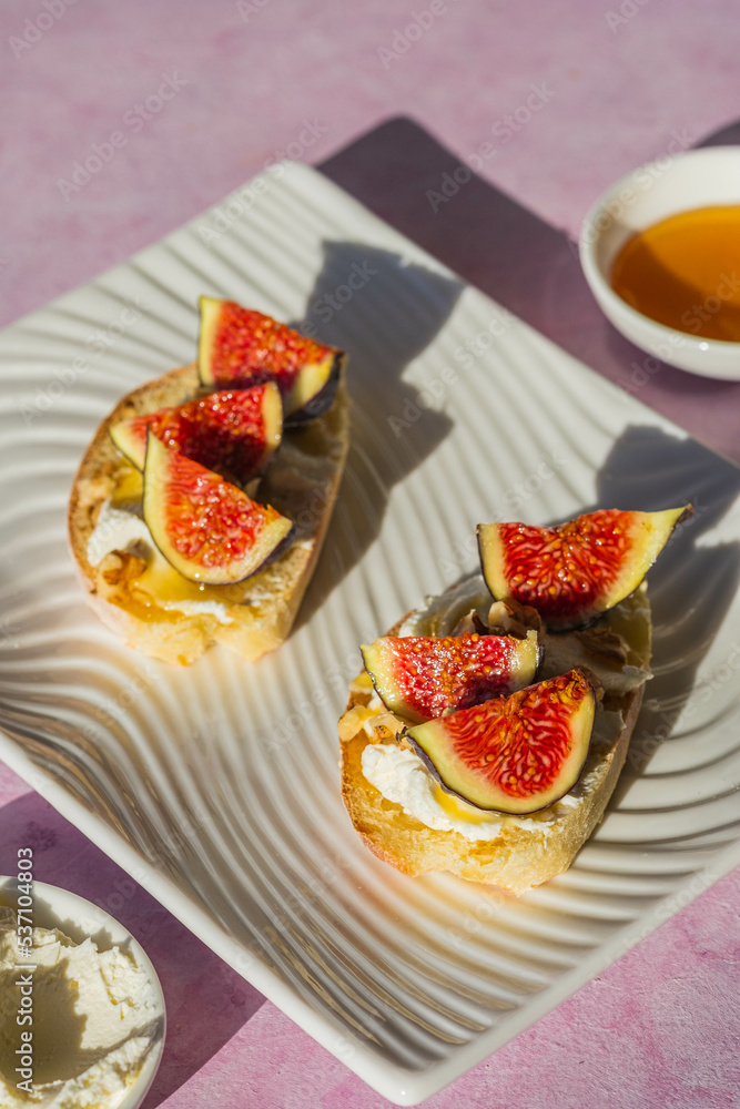 Open sandwiches or bruschetta with cream cheese, fresh figs, honey and walnuts on a white rectangular plate on a pink concrete background. Open sandwiches or bruschetta with cream cheese, fresh figs, honey and walnuts on a white rectangular plate on a pink concrete background.