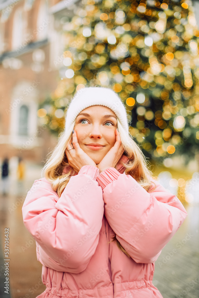 A cute blonde in a hat and a pink jacket against the background of a Christmas tree on the street. Winter walk, new year, feast day. A cute blonde in a hat and a pink jacket against the background of a Christmas tree on the street. Winter walk, new year, feast day.