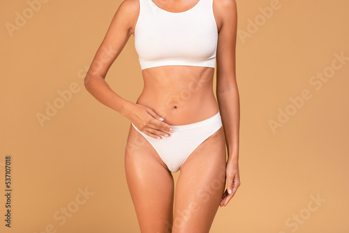 Unrecognizable well-fit woman in white underwear posing in top bra and panties over beige studio background, cropped