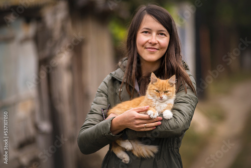 Portrait of young woman glasses holding cute red cat with green eyes. Female hugging her cute long hair kitty. Background, copy space, outdoors beautiful playing brunette.