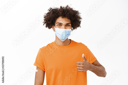 Young hispanic man recommends do covid 19 vaccine shot, join vaccination campaign, showing thumbs up, wearing medical face mask, standing over white background © Cookie Studio