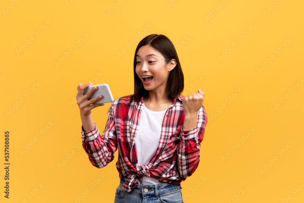 Overjoyed asian lady holding modern smartphone and clenching fist, gambling or trading online over yellow background Overjoyed asian lady holding modern smartphone and clenching fist, gambling or trading online over yellow background
