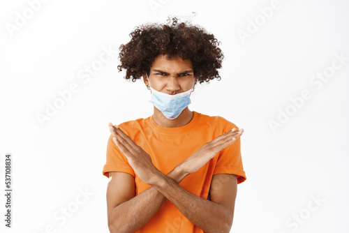Man showing stop sign, wearing medical face mask wrong, showing thumbs down, he has patch on arm after covid-19 vaccination, dislike smth, standing over white background © Cookie Studio