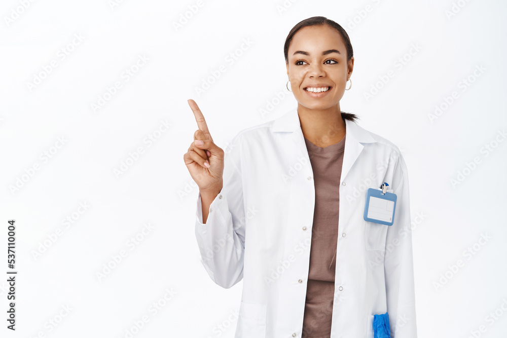 Woman doctor in white coat, pointing finger and looking at banner, showing medical clinic logo on copy space, standing over white background Woman doctor in white coat, pointing finger and looking at banner, showing medical clinic logo on copy space, standing over white background