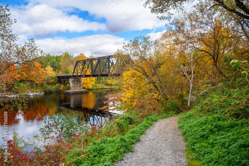 Fototapeta Naklejka Na Ścianę i Meble -  Empty rusty railroad bridge over a river on a partly cloudy autumn mornig. A riverbank path through colourful autumn trees is in foreground.