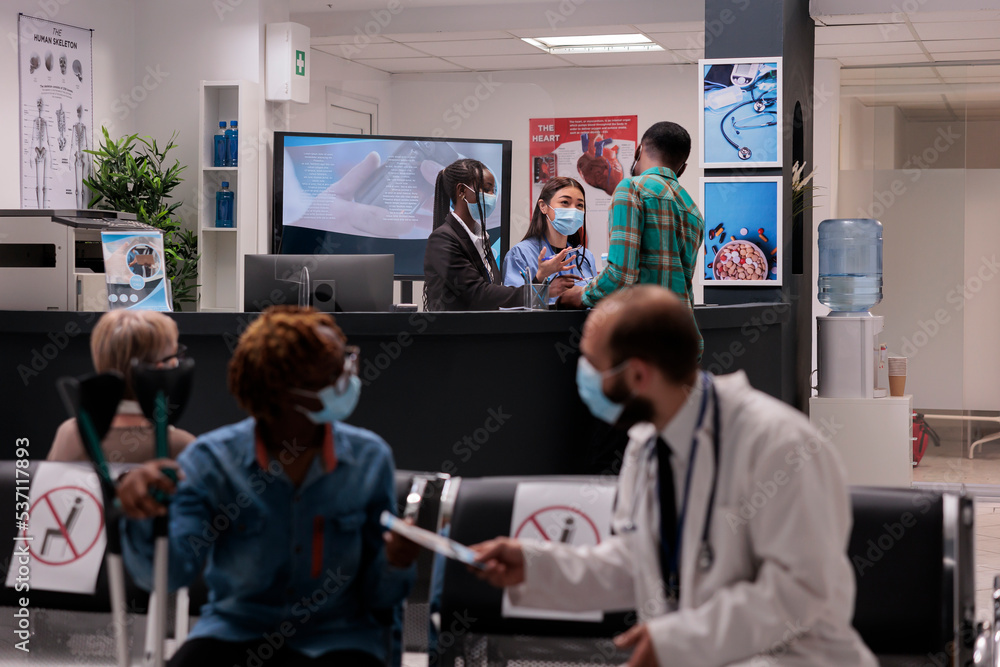 Obraz premium Female hospital staff members attending a sick young african american man at reception area. Doctor handing prescription to woman with partial body paralysis. Medical clinic lobby during covid