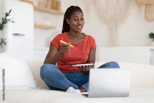 Black Young Woman Watching Webinar On Laptop At Home And Taking Notes