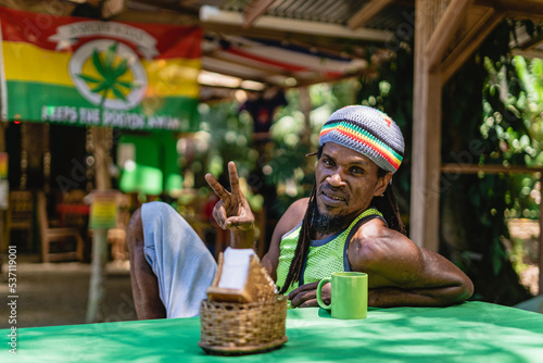 Retrato horizontal de un hombre afrocaribeño sonriente con cabello rasta sentado solo en el exterior en un día de verano haciendo un signo de paz con su mano.  photo