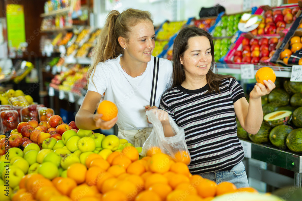 Obraz premium Young woman with a fifteen-year-old girl who came to the supermarket for shopping, choose oranges at the counter, putting ..them in a package