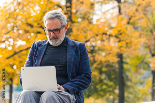 Outdoor office in city park during beautiful autumn day. Internet browsing concept. Focused 60-year-old caucasian male entrepreneur dressed in business-casual way sitting with laptop on his knees and