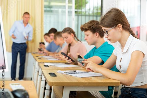 Concentrated teenage girl with group of fellow students sitting at lesson in classroom  using mobile phone