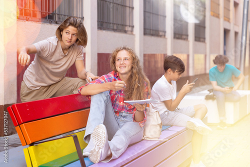 Guy and girl have fun chatting on bench. Guy points his hand at someone