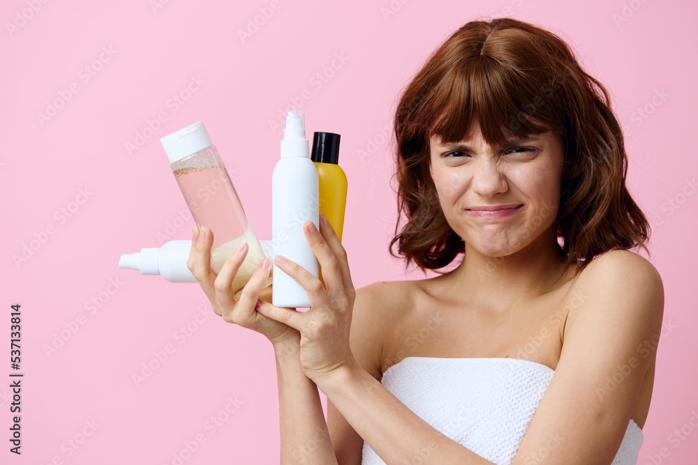 Fototapeta premium a sad, worried woman stands on a pink background, wrapped in a white towel and holding a set of jars with facial care cosmetics in her hands