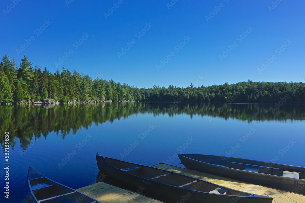 Adirondack Park, New York: Canoes tied to a wooden dock on Sagamore Lake, surrounded by evergreen trees on a bright summer day.