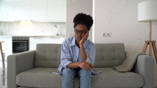 Flustered African American woman is very nervous sighing and touching face with hands sits on couch of home. Young black girl takes off glasses feeling fear after wrong decision or problems at work  photo