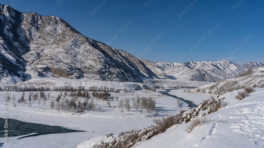 The winter River winds through the valley. Steam rises over the unfrozen part of the riverbed. Bare trees in the snow. A picturesque mountain against the blue sky. Altai. Katun