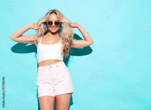 Young beautiful smiling blond female in trendy summer shorts clothes. Sexy carefree woman posing near blue wall in studio. Positive model having fun indoors. Cheerful and happy. In sunglasses