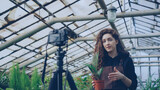 Popular female blogger experienced gardener is recording video about gardening for her vlog standing in greenhouse and holding pot flowers. Young people and botany concept.