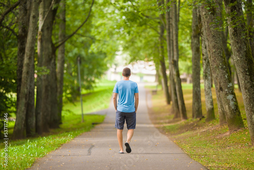 Young adult man slowly walking through alley of green trees on sidewalk at city park in warm summer day. Spending time alone. Peaceful atmosphere. Back view.
